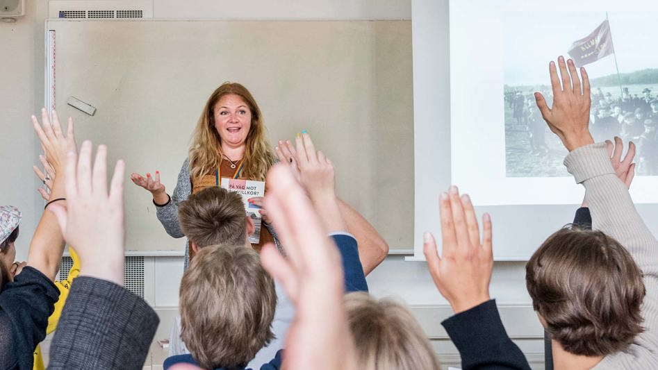En lärare står i ett klassrum fyllt med elever som räcker upp sina händer.