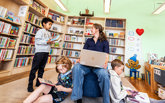 Bibliotekarie som sitter med dator i knät inne på biblioteket. Barn som tittar i böcker.