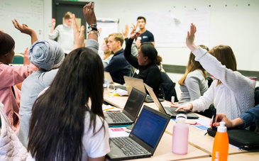 Elever sitter runt ett bord i ett klassrum, några med datorer framför sig, och räcker upp handen.