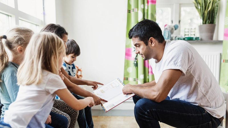 Lärare sitter på liten stol och håller upp en uppslagen bok för flera barn som pekar i den.