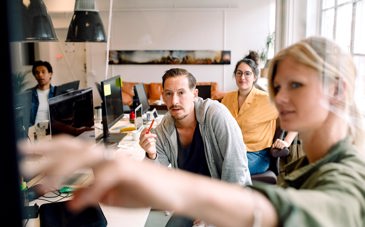 En kvinna visar något på en vägg med sin arm, medan två andra sitter och intresserat tittar på.