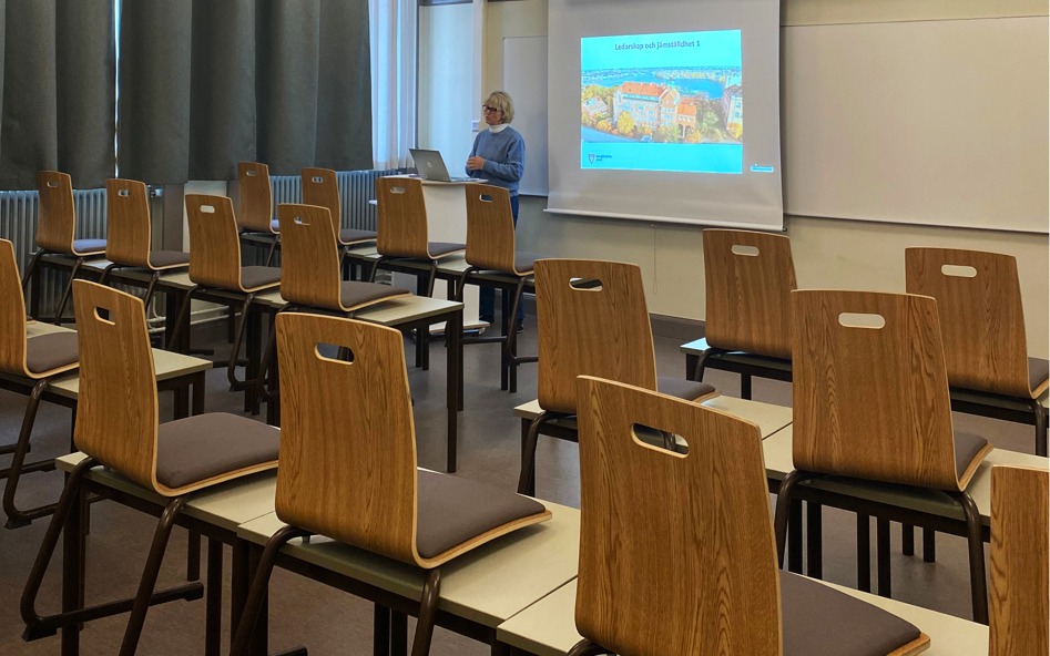 Stolar uppställda på tomma bord. Lärare undervisar framför dator.