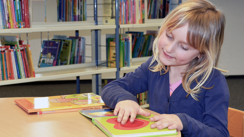 En flicka i ett biblioteket har handen på en bild i en bok. 