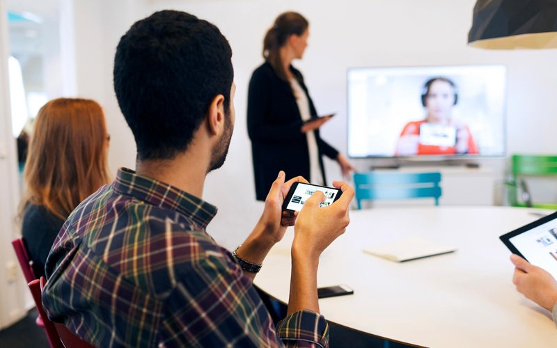 Personer sitter kring ett konferensbord och en person deltar från distans och syns på en skärm.