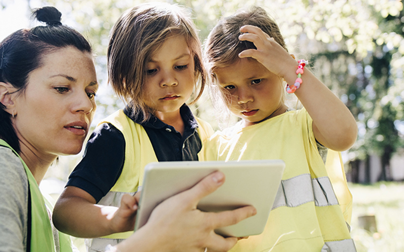 En pedagog och två förskolebarn med gula västar tittar på en ipad utomhus.