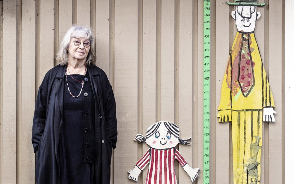 En kvinna i 70-årsåldern står lutad mot en vägg vid barnboksfigurerna Lilla Anna och Långa farbrorn.