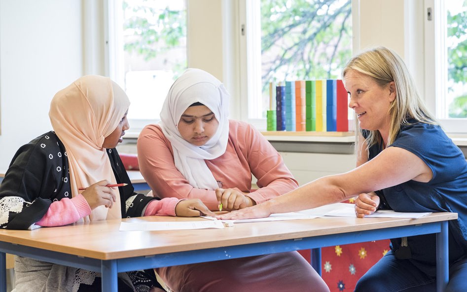En lärare och två elever vid ett bord, diskuterar ett papper på bordet som läraren pekar på.