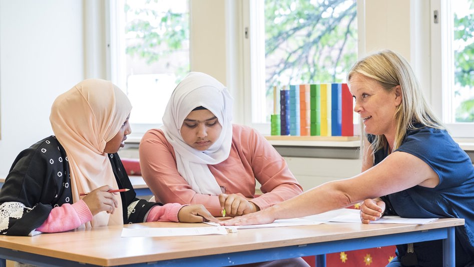En lärare och två elever vid ett bord, diskuterar ett papper på bordet som läraren pekar på.