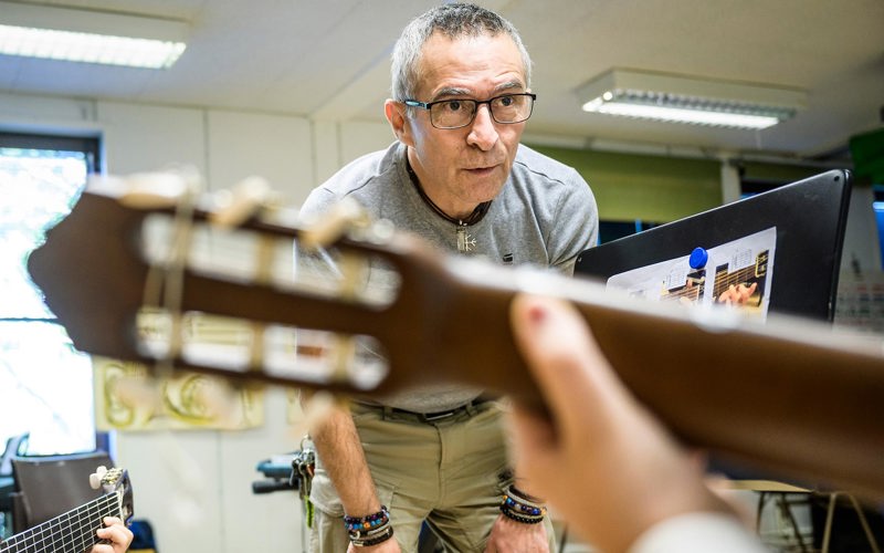 En lärare står framåtlutad med positiv och koncentrerad blick på någon som spelar gitarr.