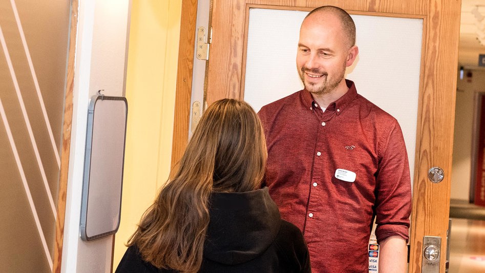 En man i röd skjorta står vid klassrumsdörren och välkomnar en av sina elever med ett leende.