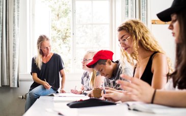Elever i gymnsieåldern sitter vid ett bord och läser intensivt. En lärare sitter på bordet.