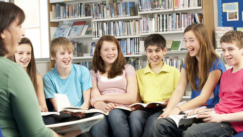 Elever med uppslagna böcker i knät sitter på stolar runt en pedagog i biblioteket.