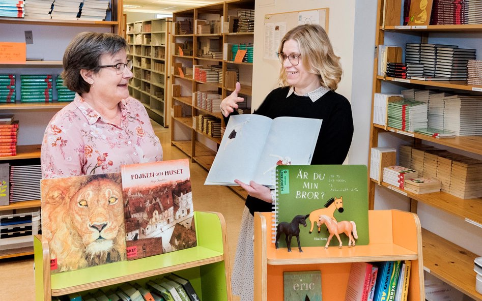 Två kvinnor står i en biblioteksmiljö och håller upp böcker.