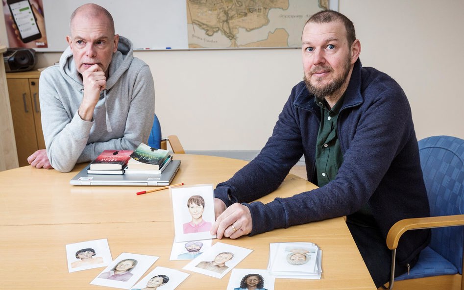 Två medelålders män sitter vid ett konferensbord. Framför dem ligger tecknade bilder av människor.