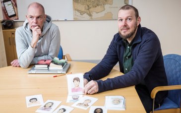 Två medelålders män sitter vid ett konferensbord. Framför dem ligger tecknade bilder av människor.