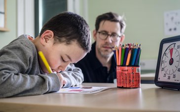 En elev sitter vid bord och tecknar koncentrerat medan en lärare sitter i bakgrunden.