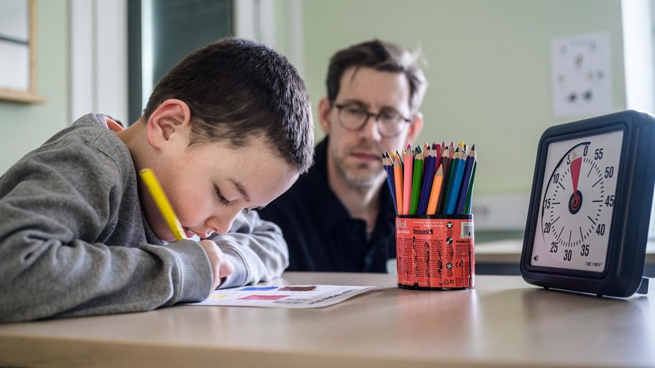 En elev sitter vid bord och tecknar koncentrerat medan en lärare sitter i bakgrunden.
