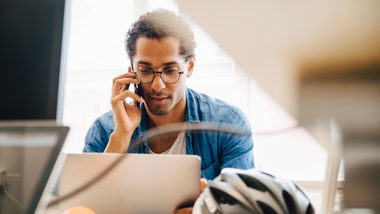 En man sitter vid en dator och talar i mobiltelefon.