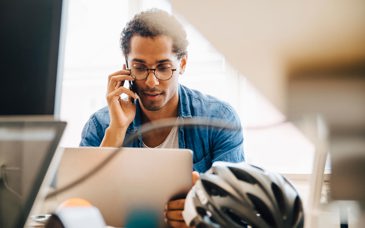 En man sitter vid en dator och talar i mobiltelefon.