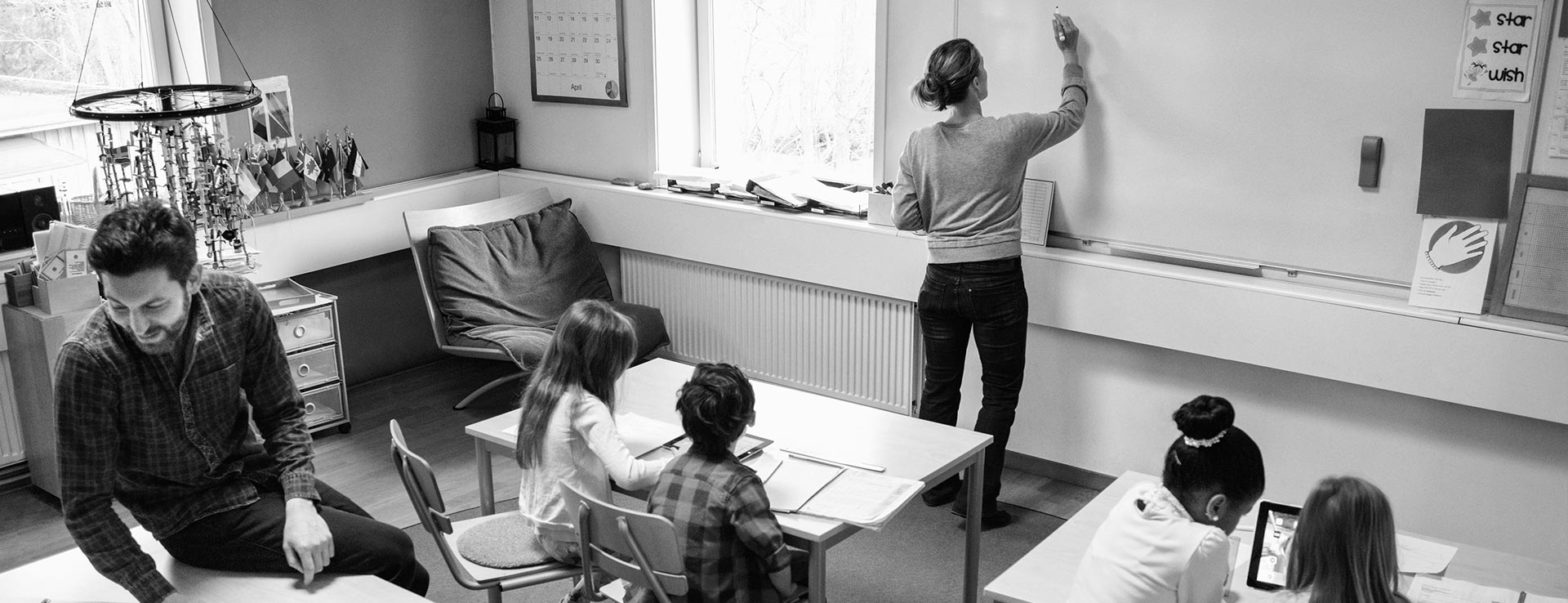 Ett fullsatt klassrum med en lärare som skriver något på tavlan och en annan pratar med en elev.