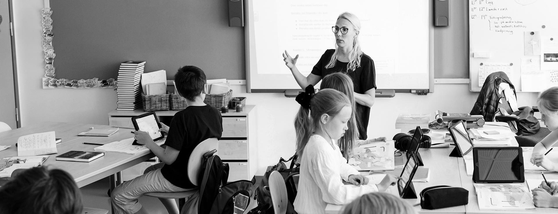 En lärare i ett klassrum gestikulerar med händerna. Kring henne sitter elever med surfplattor.