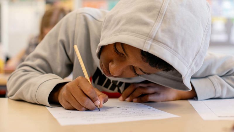 En elev sitter i ett klassrum och skriver med papper och penna 
