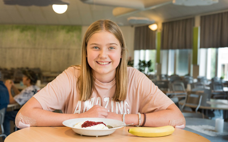 En leende elev sitter vid ett bord med en tallrik mat och en frukt.