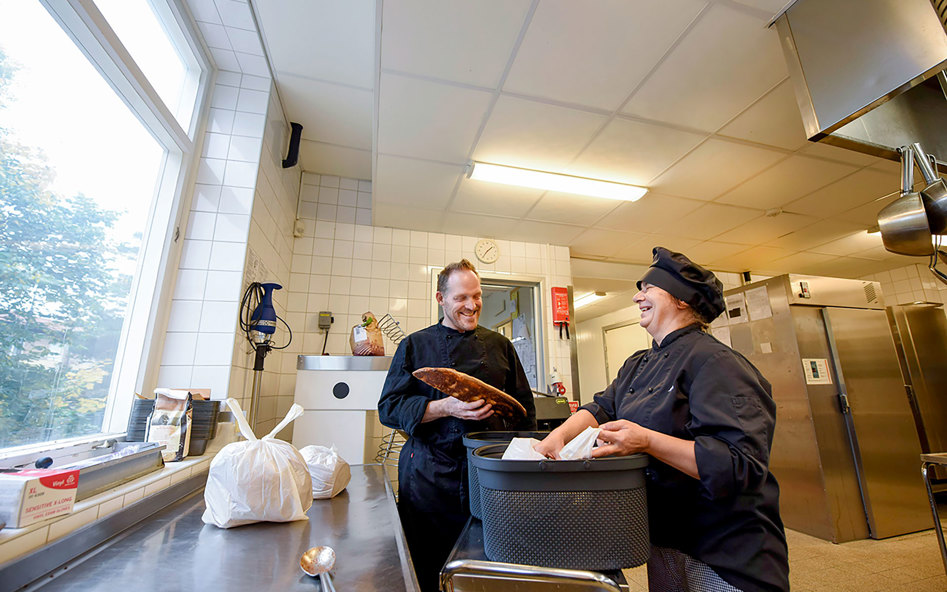 En man med ett surdegsbröd i handen samtalar med en kvinna. De står i ett skolkök.
