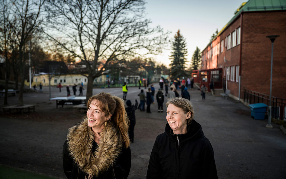 Två leende kvinnor på en skolgård. I bakgrunden syns elever, träd och skolbyggnader.