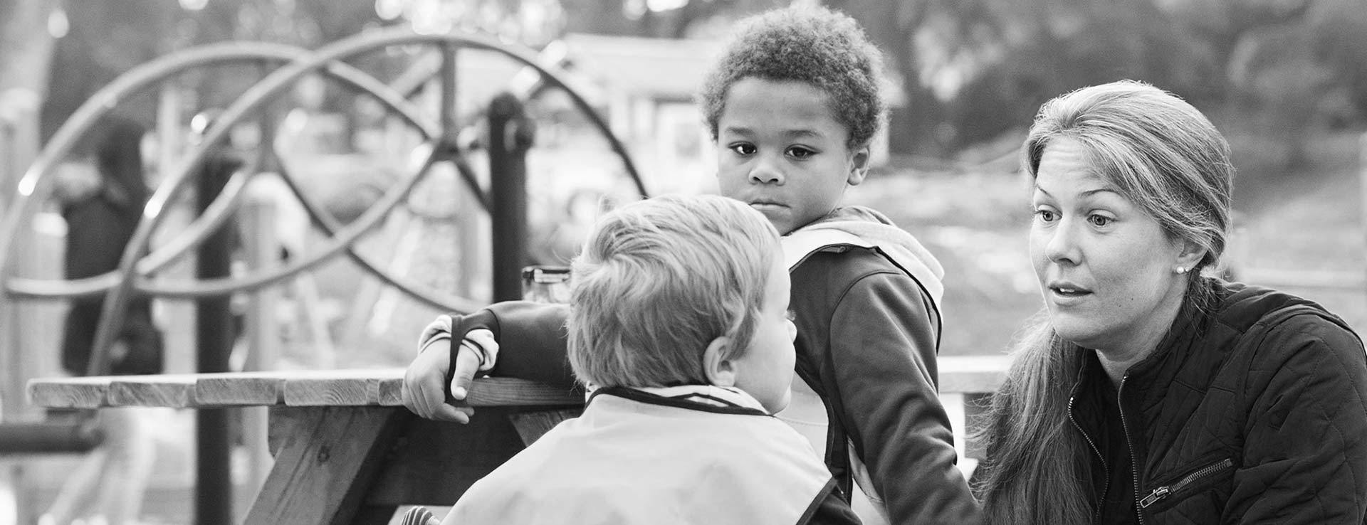 En förskolepedagog pratar med två förskolebarn på en lekplats.