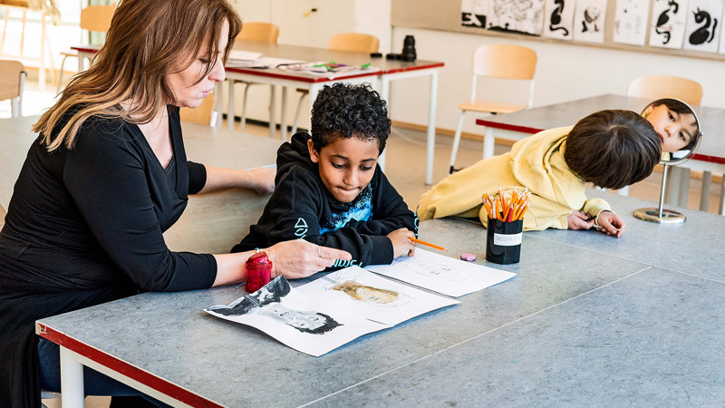 Lärare och två elever i bildsal. De tittar på den ena elevens porträtt.