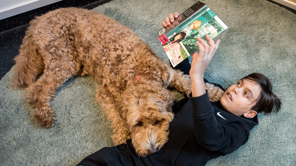 Pojke ligger på golv och läser en bok. Hund ligger bredvid med huvudet på pojkens mage.