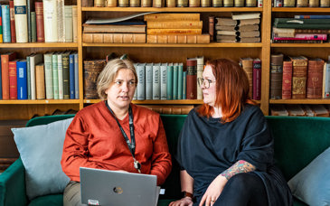 Två kvinnor sitter i en soffa framför en stor bokhylla. De ser ut att samtala. 