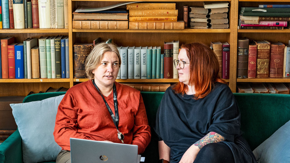 Två kvinnor sitter i en soffa framför en stor bokhylla. De ser ut att samtala. 