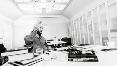 En man sitter vid ett skrivbord med böcker. Han pratar och pekar med handen.