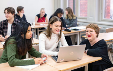 En kvinnlig lärare sitter på huk bredvid två gymnasieelever och tittar på en datorskärm.