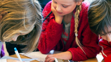 Tre barn som skriver, ritar och läser vid ett bord.