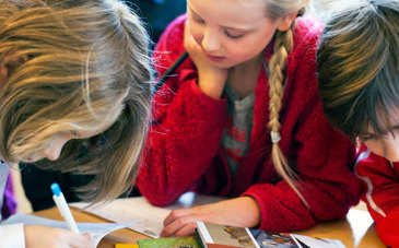 Tre barn som skriver, ritar och läser vid ett bord.