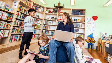 Bibliotekarie som sitter med dator i knät inne på biblioteket. Barn som tittar i böcker.