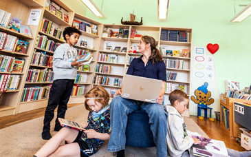 Bibliotekarie som sitter med dator i knät inne på biblioteket. Barn som tittar i böcker.