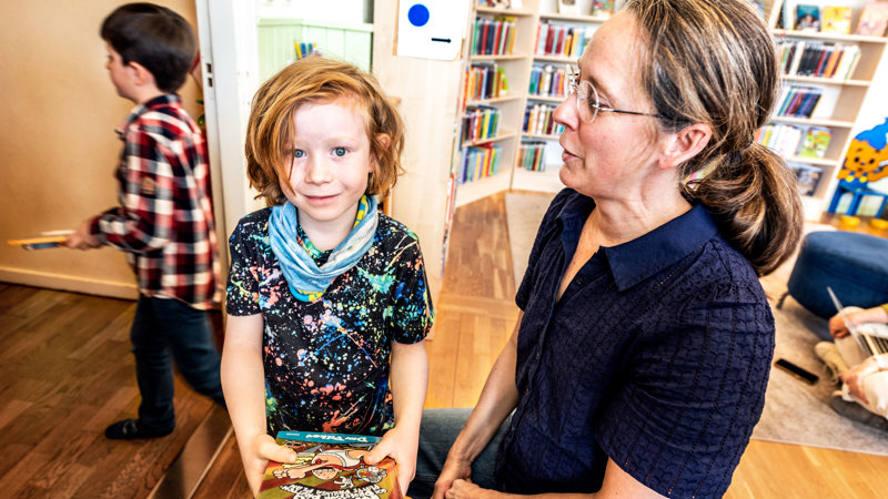 Barn som håller i bok. Bibliotekarie som sitter på huk bredvid barnet.