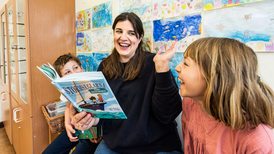 En lärare och två elever läser ur en bok och skrattar.