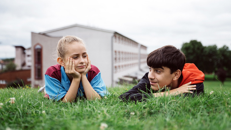 En flicka och en pojke ligger på mage på gräsmattan och tittar på varandra.
