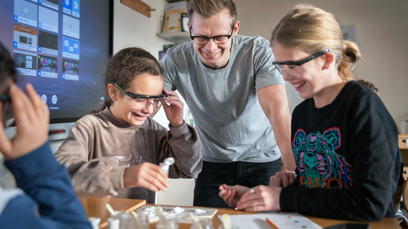 Två elever med skyddsglasögon experimenterar och en pedagog tittar på.