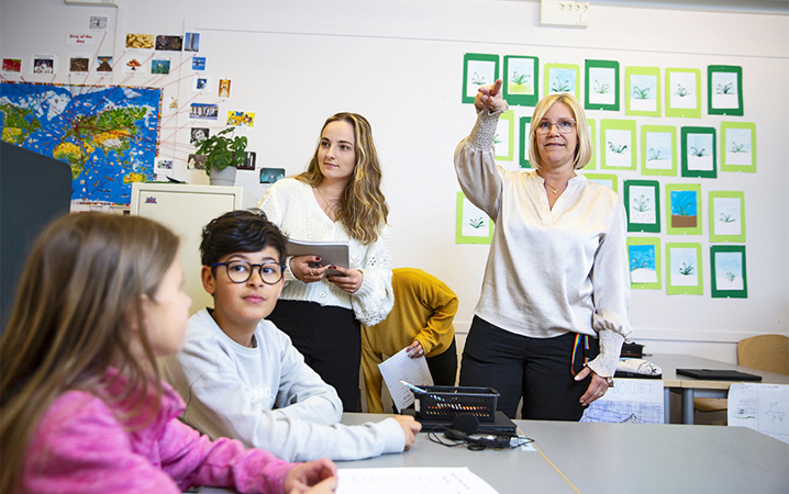 Fyra elever och en lärare i ett klassrum. Läraren pekar på något med handen.