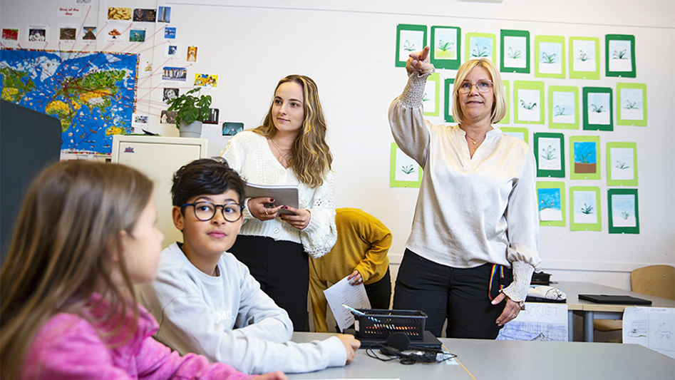 Fyra elever och en lärare i ett klassrum. Läraren pekar på något med handen.