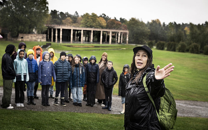 En grupp med barn ute på ett fält tittar på en kvinna som pekar på något