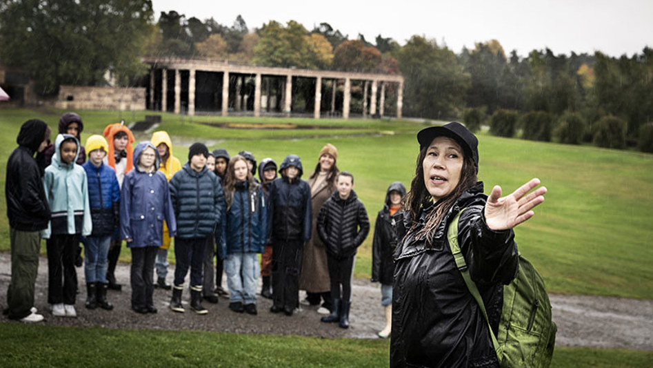 En grupp med barn ute på ett fält tittar på en kvinna som pekar på något