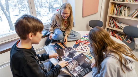Tre glada elever med uppslagna böcker på ett runt bord i ett bibliotek.