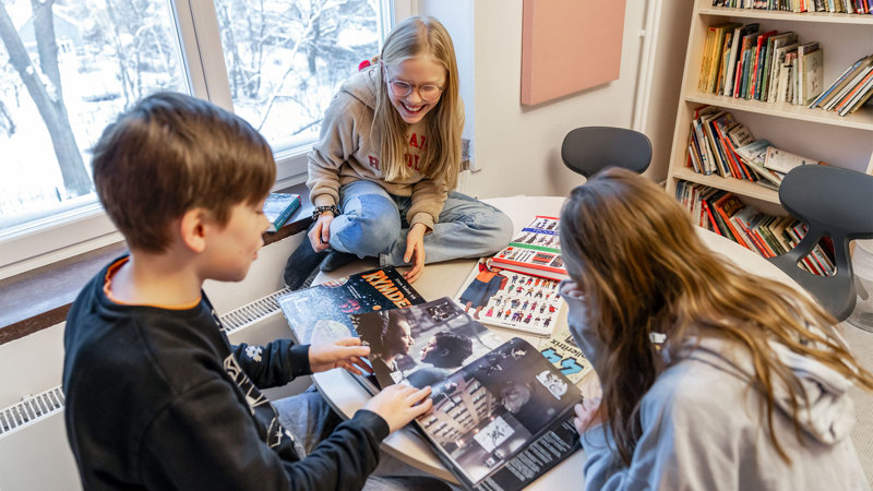 Tre glada elever med uppslagna böcker på ett runt bord i ett bibliotek.